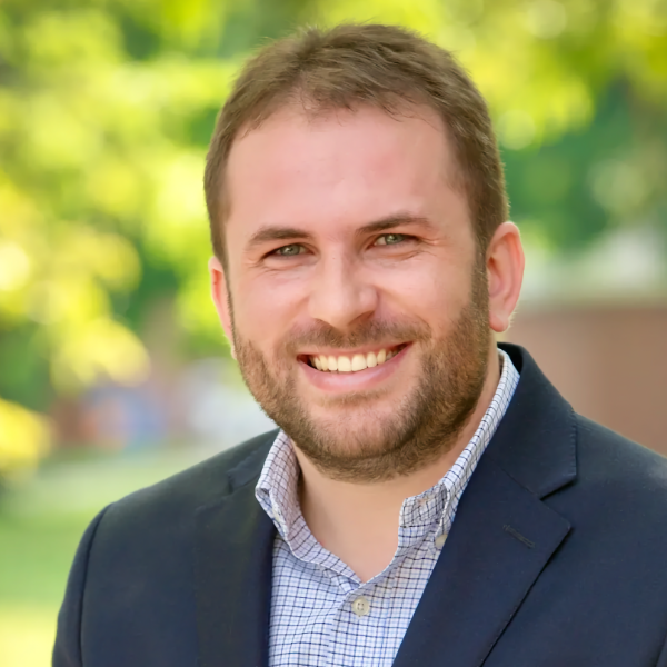 Professional portrait of Dr. Ribeiro smiling, wearing a navy blue blazer and a checkered shirt, with a blurred outdoor greenery background.