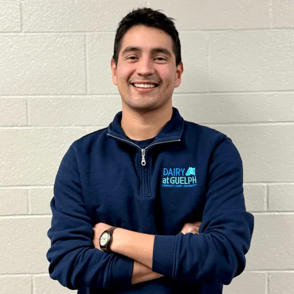 A man smiling with his arms crossed, wearing a navy blue quarter-zip pullover with a "Dairy at Guelph" logo, posing against a white brick wall.