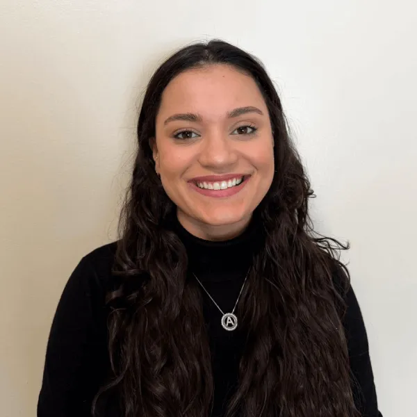 A portrait of a smiling woman with long, dark wavy hair, wearing a black turtleneck sweater and a silver initial necklace against a plain off-white background.
