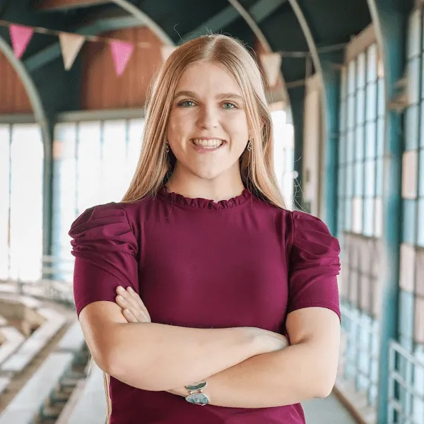 A smiling woman with long blonde hair, posing with her arms crossed and wearing a burgundy short-sleeved blouse in an airy, industrial-style building.