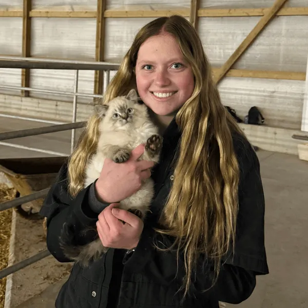 A smiling young woman with long blonde hair holding a fluffy grey and white kitten, wearing a black work jacket inside a farm building.