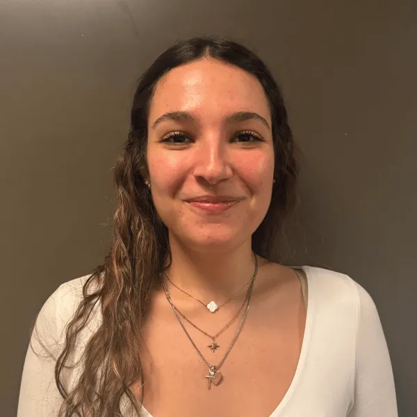 A portrait of a smiling woman with long curly brown hair, wearing a white long-sleeved top and layered gold necklaces against a dark grey background.