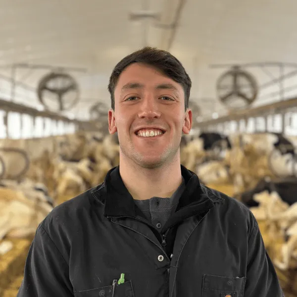 A portrait of a man smiling with short dark hair, wearing a black work jacket over a grey shirt. He is standing in a large dairy barn with rows of cows and industrial fans visible in the blurred background.