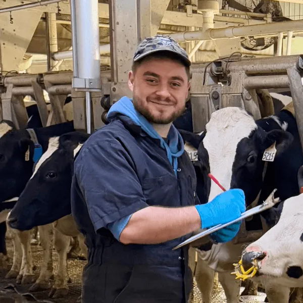 A smiling man wearing a camouflage cap, blue hoodie, and black overalls, holding a clipboard and pen while standing next to Holstein cows in a milking parlor.