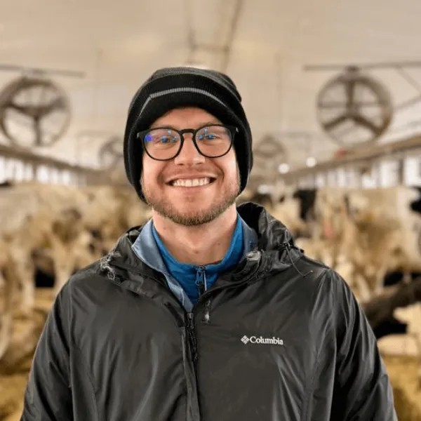 A portrait of a smiling man with glasses and a black beanie, wearing a black Columbia jacket inside a dairy facility with cattle in the background.
