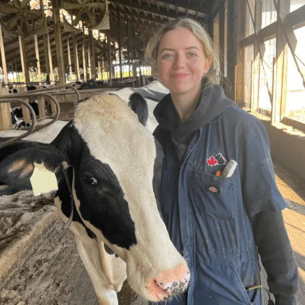 A woman smiling while standing next to a Holstein cow inside a barn, wearing navy blue coveralls with a red maple leaf logo.