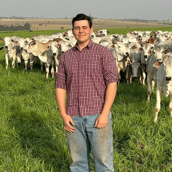 A portrait of a man in a maroon and white checkered shirt and blue jeans, standing in a lush green pasture. Behind him, a large herd of Nelore cattle is gathered. The background shows a vast, rolling landscape under a clear sky.