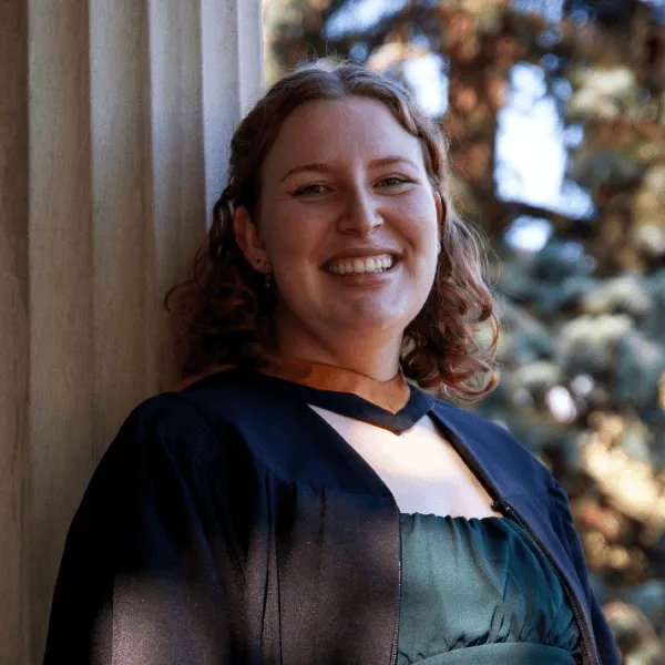 A portrait of a woman smiling with wavy reddish-brown hair, wearing a dark graduation gown over a forest green dress. She is leaning against a white classical-style pillar, with soft sunlight and blurred evergreen trees in the background.
