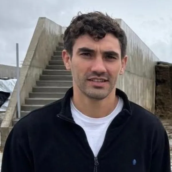 A portrait of a man with short, curly brown hair and light facial hair, wearing a black zip-up jacket over a white t-shirt. He is looking at the camera. He is outdoors in a farm setting, with a concrete bunker silo containing dark feed and concrete stairs in the background under a cloudy sky.