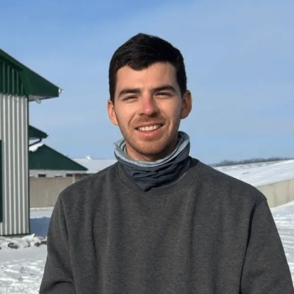 A portrait of a man smiling with short dark hair, wearing a grey sweatshirt and a grey neck gaiter. He is standing outdoors in a snowy landscape with a clear blue sky and a farm building with a green roof in the background.
