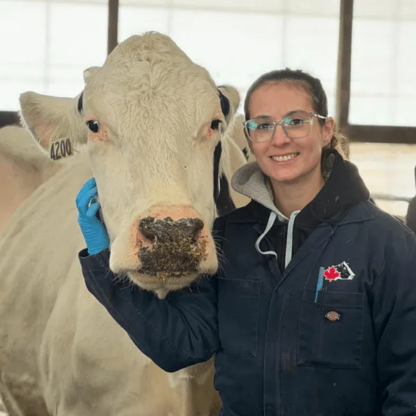 A portrait of a woman smiling with glasses, wearing a navy blue work jumpsuit with a Canadian flag patch over a grey hoodie. She has one gloved hand resting on the side of a large white cow's face. They are inside a bright barn with a white tarp wall in the background.