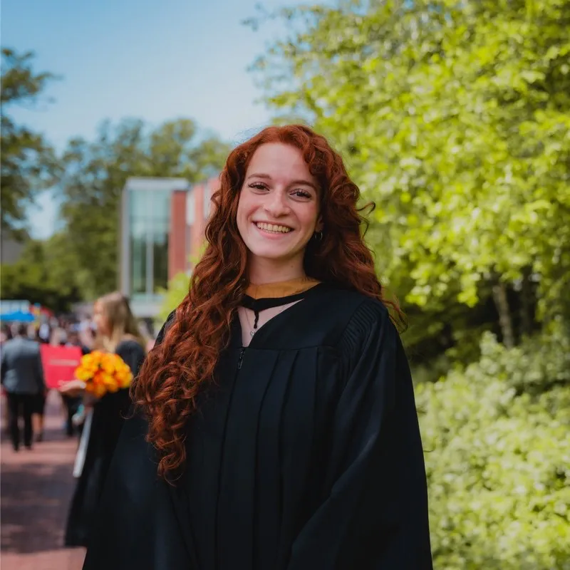 A smiling woman with long, curly red hair wearing a black academic graduation gown, posing outdoors on a university campus with green trees and buildings in the background.