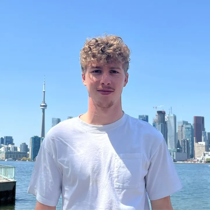 A portrait of a man with curly blonde hair wearing a white t-shirt, posing in front of the Toronto skyline and waterfront under a clear blue sky.