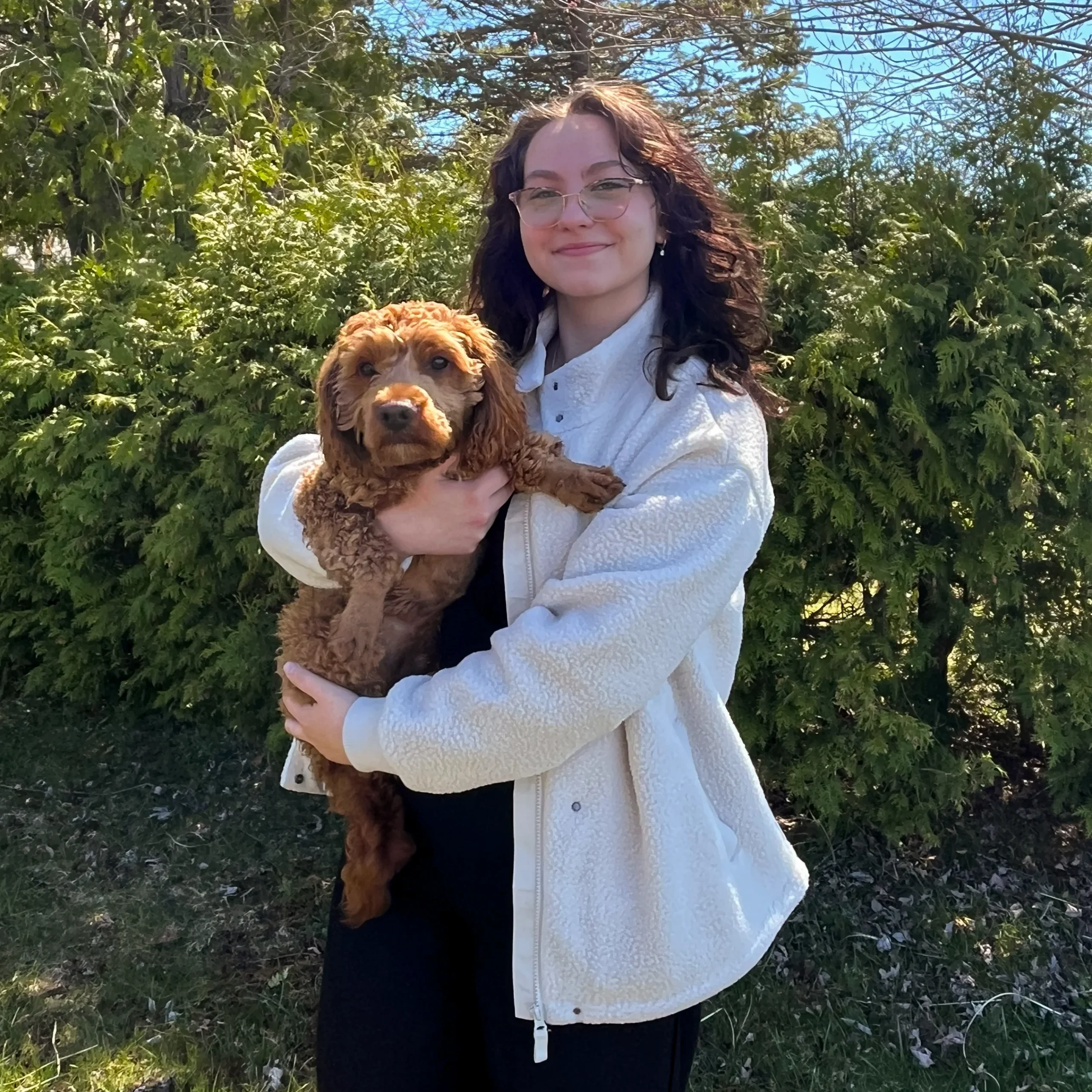 A smiling woman with dark wavy hair and glasses holding a brown curly-haired dog outdoors, with green bushes and trees in the background.