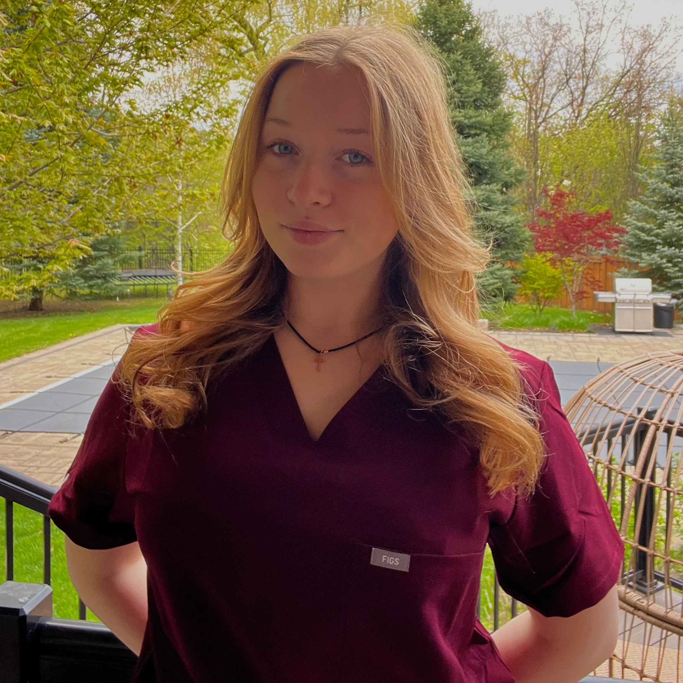 Professional portrait of a woman with long wavy blonde hair wearing maroon scrubs, standing outdoors with trees and a patio in the background.