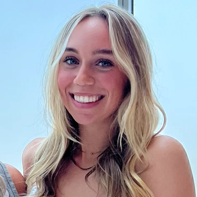 Professional portrait of a smiling woman with long blonde wavy hair, looking forward against a light background.