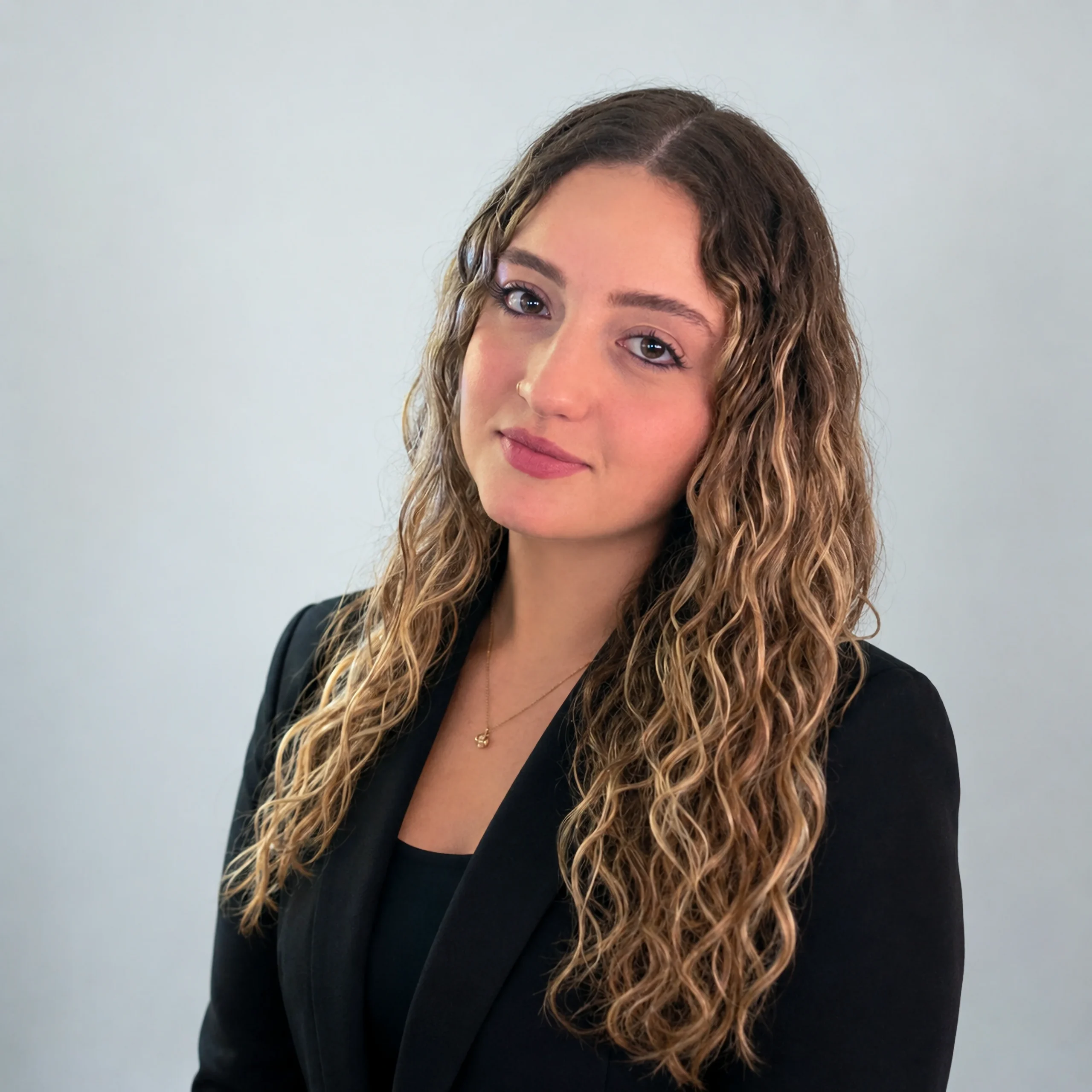 Professional portrait of a woman with long, curly light brown hair wearing a black blazer and a delicate gold necklace, smiling slightly against a grey background.