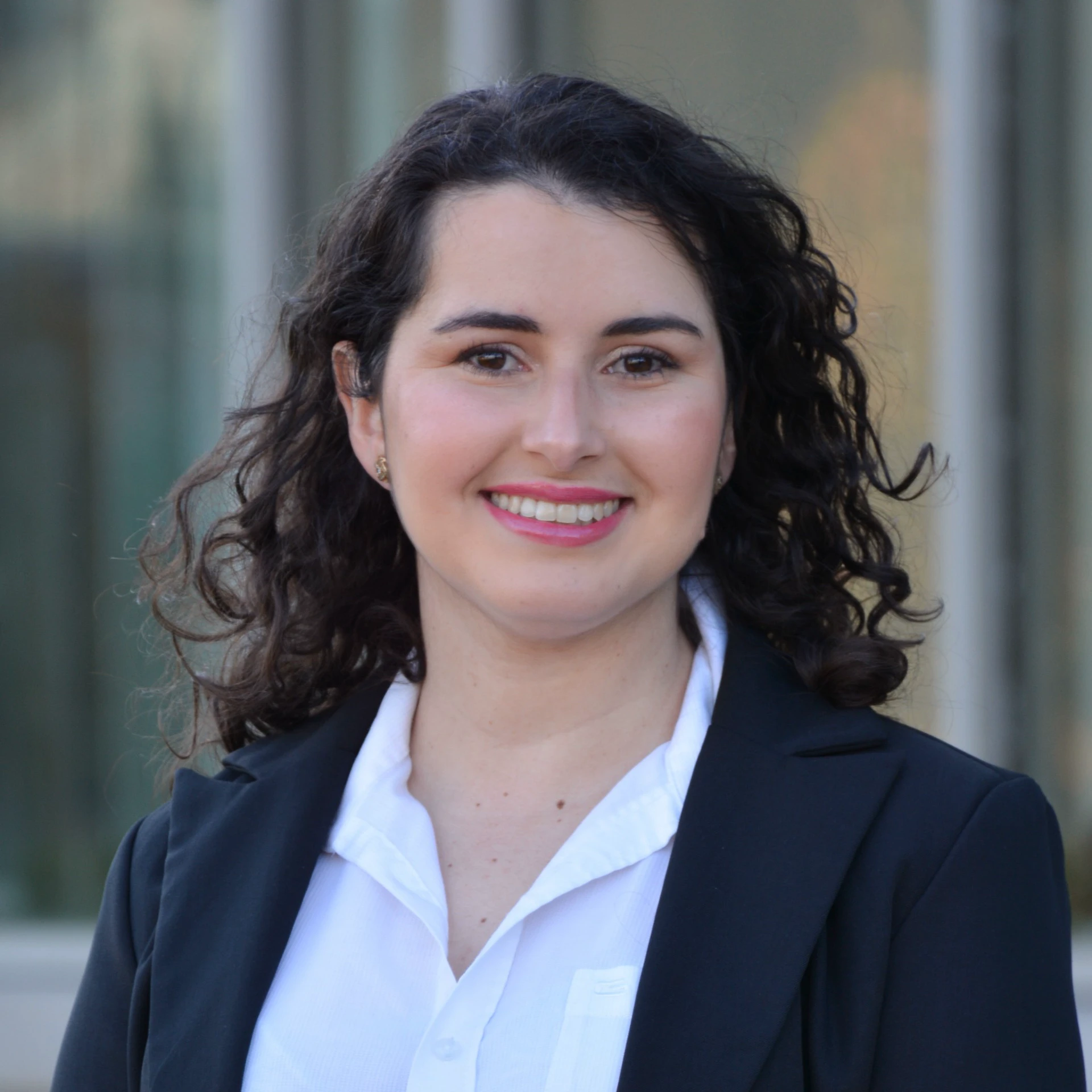 Professional portrait of a smiling woman with long curly dark hair wearing a white collared shirt and a black blazer, standing outdoors with a blurred building facade in the background.