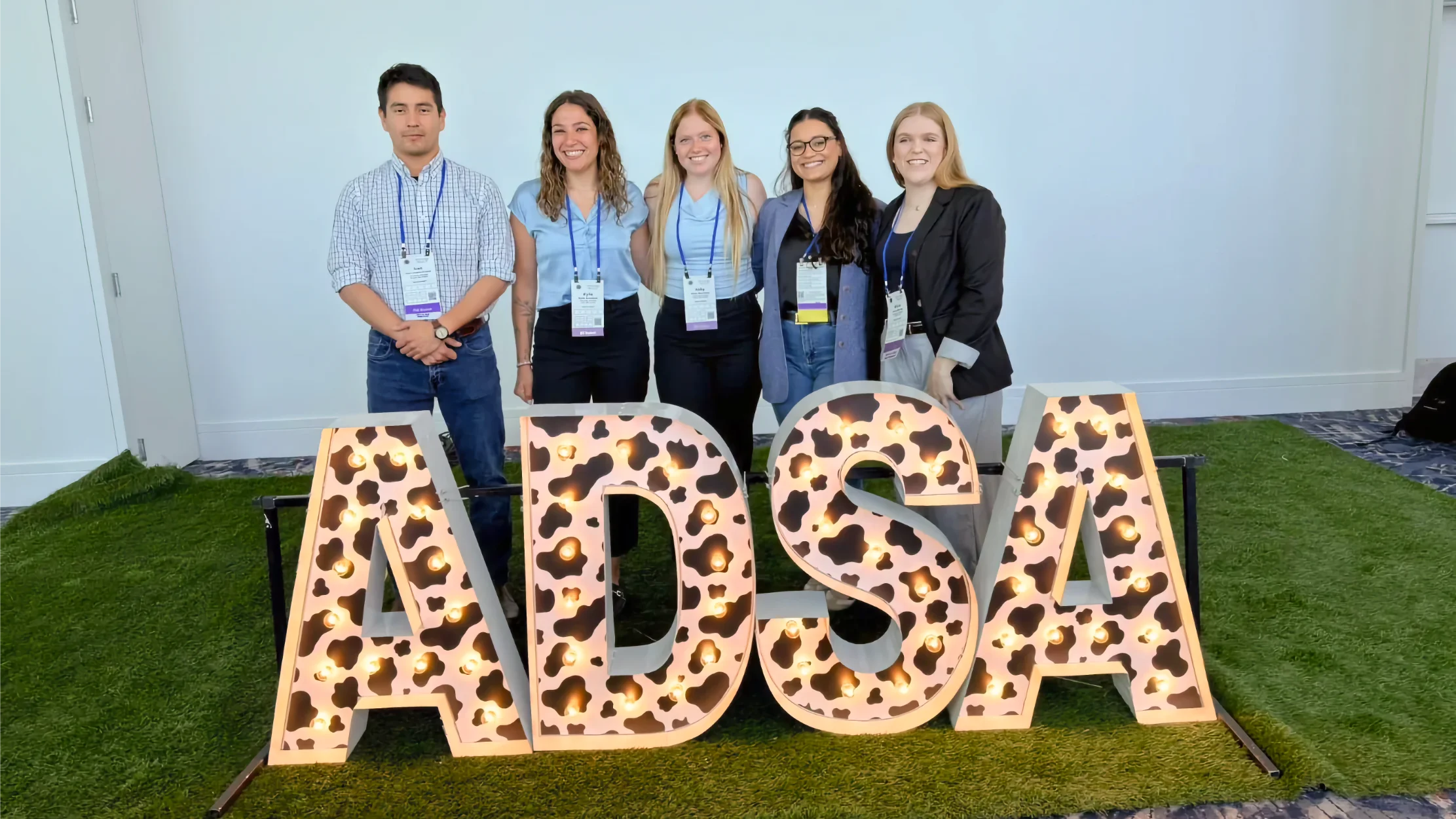 Ribeiro Lab at ADSA 2026 Wrap-Up Five researchers standing together and smiling behind large, illuminated marquee letters that spell 'ADSA' decorated with a black-and-white cow print pattern, set on an artificial grass turf background at a conference venue.