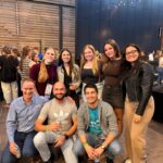 A group of eight researchers smiling and posing together at an evening social event during a conference. Three individuals are kneeling in the front row, and five are standing behind them against a modern wood-paneled and dark metal architectural wall decorated with string lights.