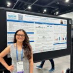 A smiling female PhD student with glasses standing with her hand on her hip next to her research poster at a conference. The poster features University of Guelph and Ribeiro Lab branding, titled 'Growth differentiation factor-15 as a novel marker for systemic inflammation in transition dairy cows'.