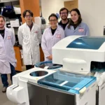 Five researchers wearing white lab coats and visitor badges stand behind a Cobas clinical chemistry analyzer during a site visit to the Animal Health Lab.