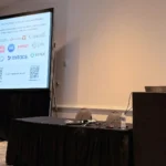 A male researcher presenting at a podium microphone during a scientific conference. The slide on the large projection screen behind him acknowledges research grants and partners, displaying institutional logos including the University of Guelph, Ontario, Canada, NSERC, Mitacs, Merck, and the Ribeiro Lab.
