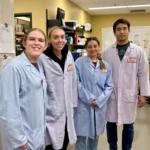 Four university researchers wearing light blue laboratory coats and visitor badges pose for a photo inside the Animal Health Lab, with research equipment and a radioactive work area visible in the background.