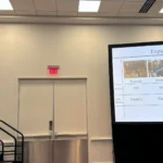 A female researcher giving an oral presentation next to a large projection screen titled 'Expanding the Data Set'. The screen displays a data table detailing cow numbers and milk testing frequencies across four different farms (Farm A1, Farm A2, Farm B, and Farm C), accompanied by small reference photographs.