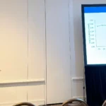 A male researcher in a blue suit gesturing with his hand while speaking at a presentation podium. The large screen behind him shows an 'Introduction' slide featuring a scientific line graph that tracks 'Total Ca in serum, mmol/L' across a timeline of days relative to calving.