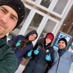 A low-angle selfie of four smiling researchers in winter clothing and blue gloves giving thumbs-up gestures during a snowfall, standing in front of a rustic wooden barn facility.