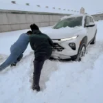 Two researchers seen from behind pushing a white SUV stuck in deep snow banks outside a dairy barn facility during a heavy winter snowfall.