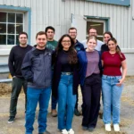 A group portrait of nine smiling researchers standing outdoors in front of a light grey barn facility with blue window frames, next to a stacked pallet of softwood shavings.