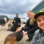 Three smiling university researchers posing for an outdoor selfie while interacting with several dogs, including a Basset Hound, a Rough Collie, and a Bernese Mountain Dog, in front of a farm building.