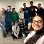 A wide-angle group selfie of eleven smiling individuals posing together indoors during a social gathering, with a close-up of a woman with long curly dark hair and glasses in the foreground, seven people standing behind her, and three people seated around a small coffee table.