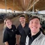 Three smiling veterinary researchers posing for a selfie inside a large, modern free-stall dairy barn with long rows of Holstein cows on both sides.