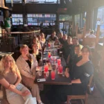 A large group of university researchers and students smiling around a long restaurant table during a social gathering, with bright sunlight streaming through the windows.