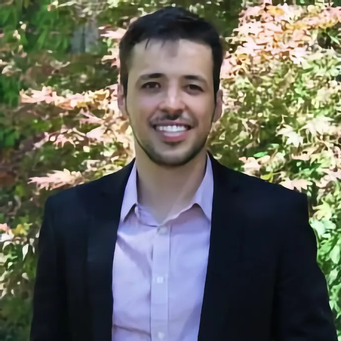 A portrait of a smiling man with short dark hair and a light beard, wearing a light pink button-down shirt and a black blazer, with a background of green and reddish foliage.
