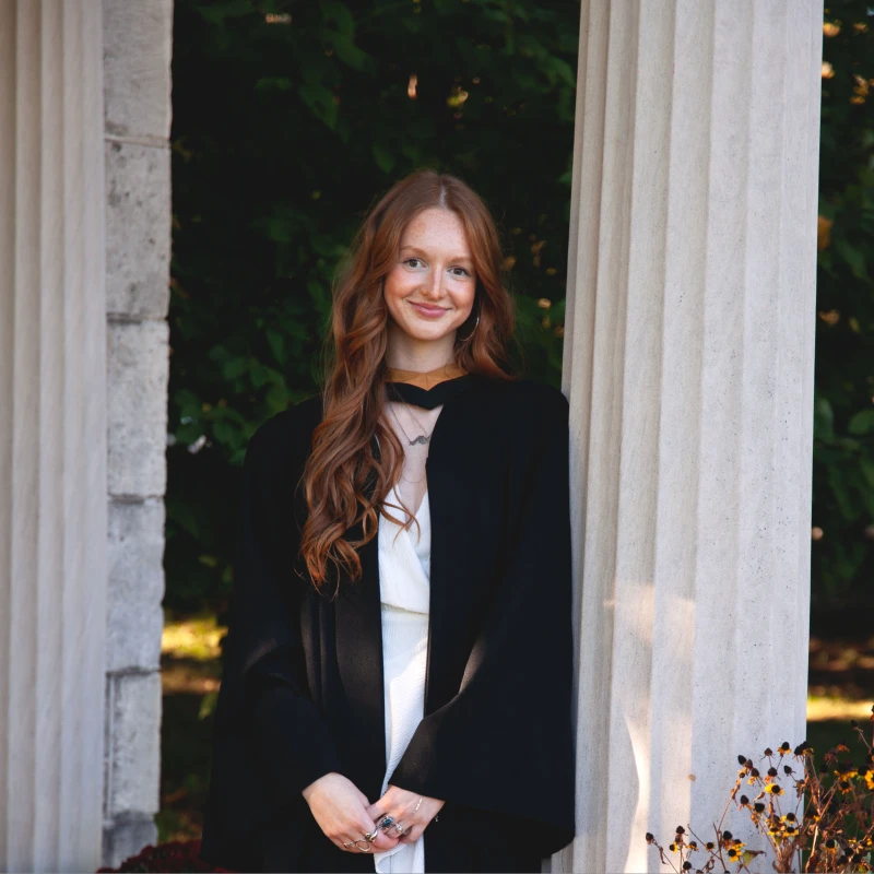 A smiling woman with long wavy red hair wearing academic graduation robes over a white dress, leaning against a classical stone pillar outdoors.