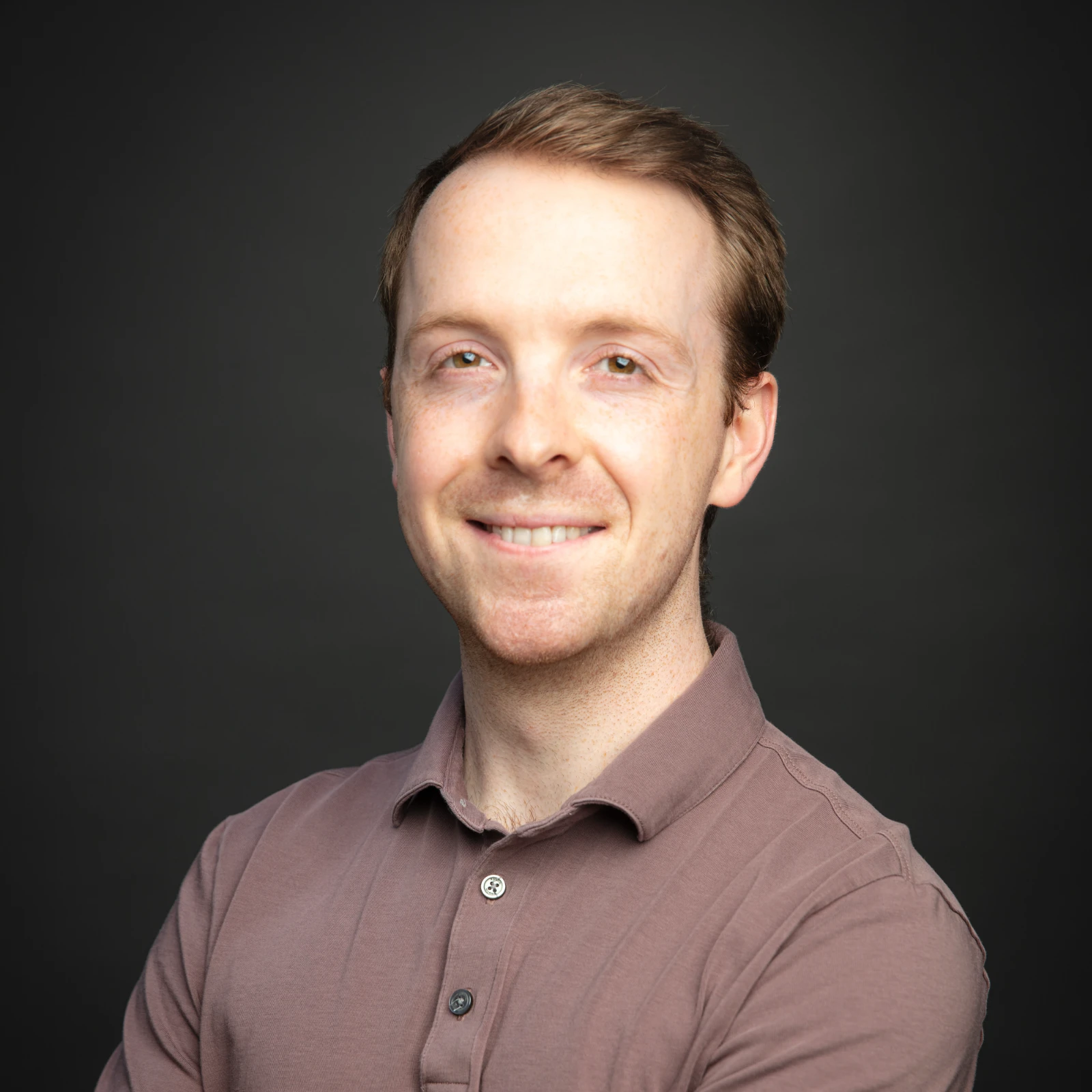 Professional headshot of a smiling young male researcher with short brown hair and light skin, wearing a mauve polo shirt, posed against a dark grey background.