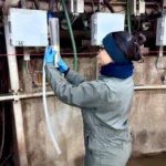 Side view of a female researcher in a grey jumpsuit and blue gloves holding up a transparent graduated cylinder containing a fresh milk sample in front of automated systems inside a dairy facility.