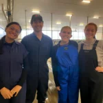 Four smiling researchers standing side by side inside a brightly lit dairy cattle barn, wearing protective farm workwear including coveralls and overalls.